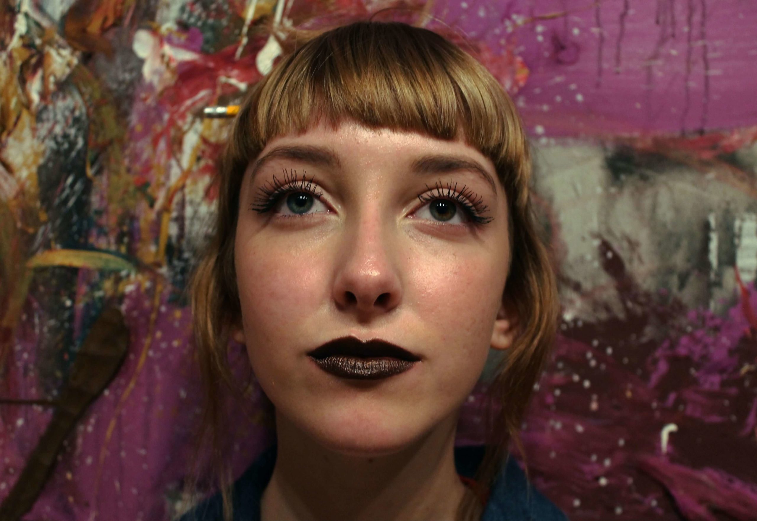 Young person with dark lipstick and blond hair in front of a painting looking at the camera