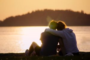 Two people cuddling by a lake at sunset.