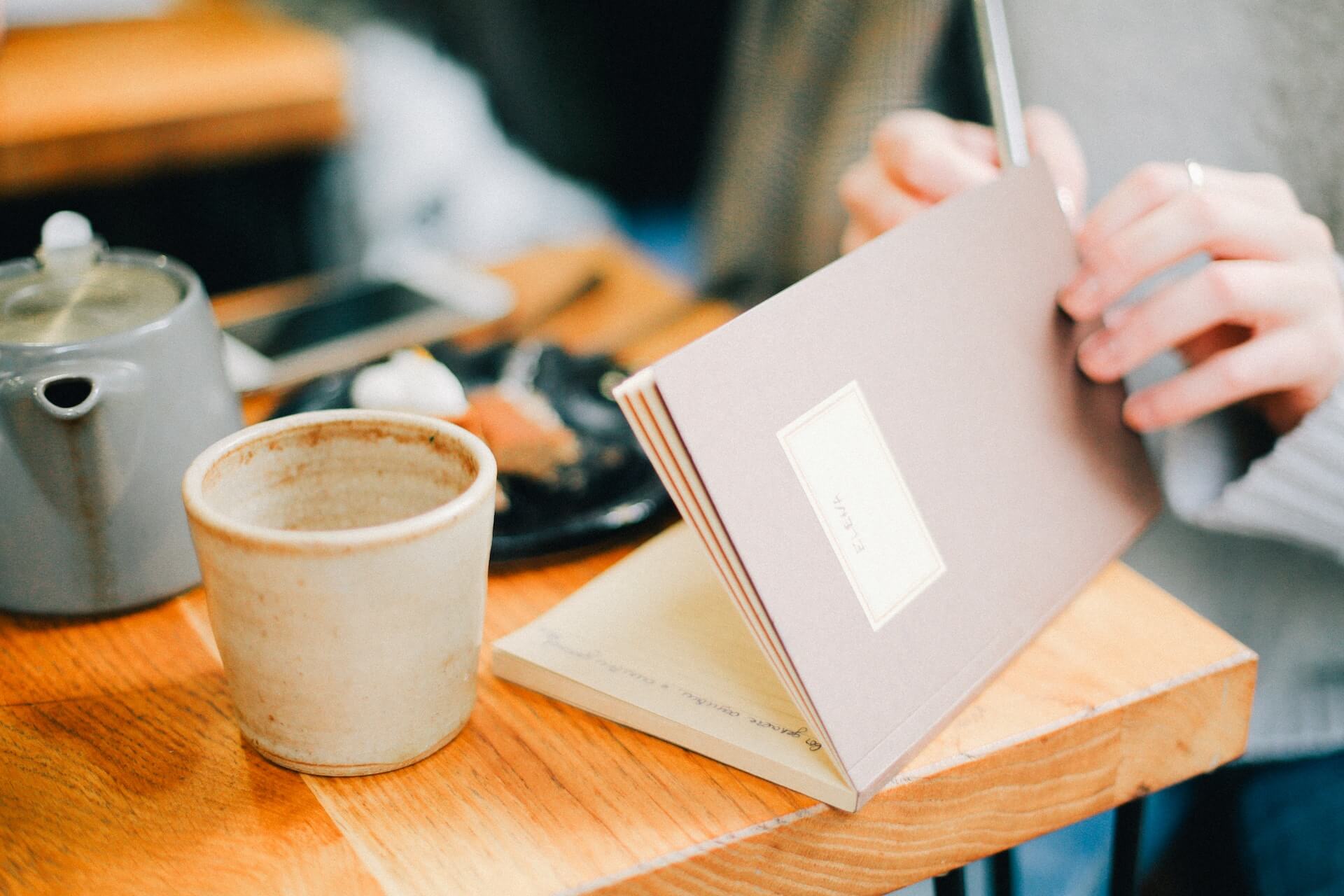 Photo of a person sitting at a cafe with a teapot, opening their journal to start writing