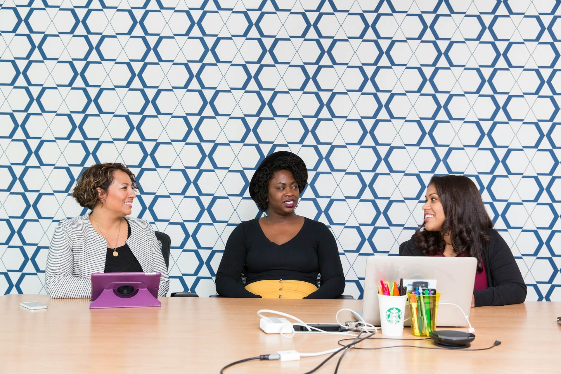 A group of 3 colleagues in a workplace comfortably chatting with each other about a project at a table with laptops and tablets on it.