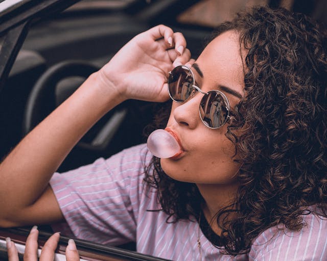 A confident woman in sunglasses blows a bubble with her chewing gum