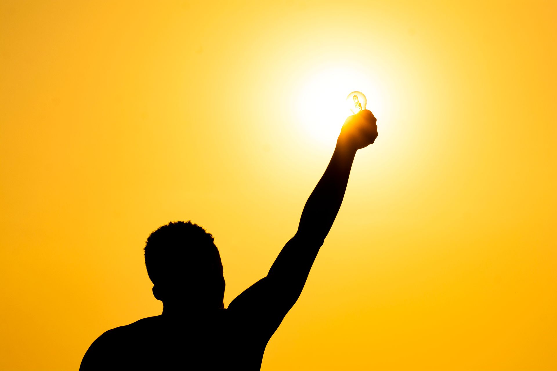 A silhouetted man holds a light bulb before a bright sun against an orange sky
